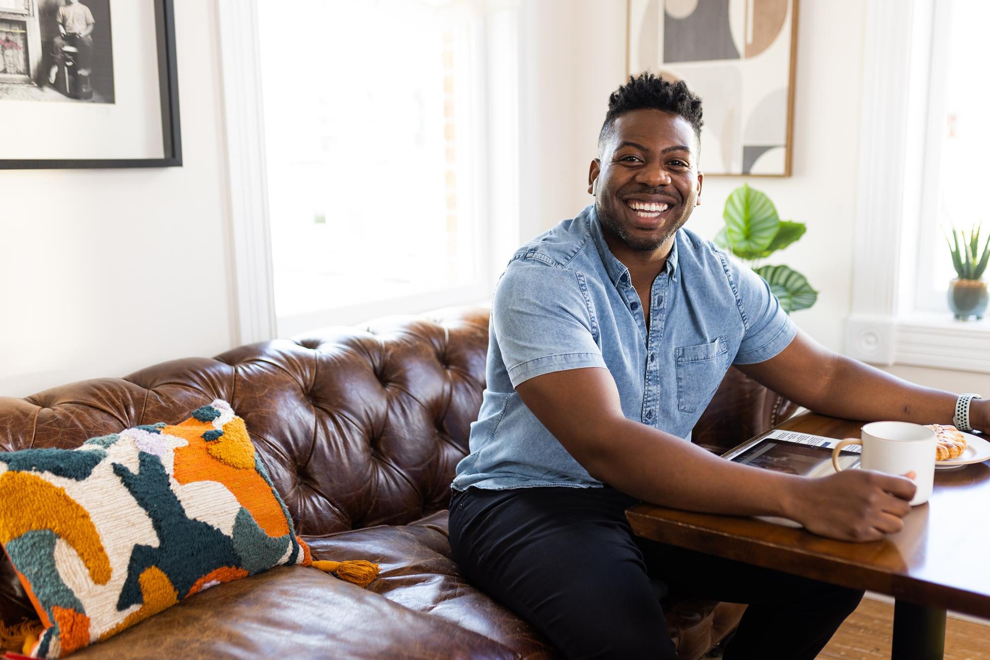 Man smiles at the camera while sitting on sofa at a table with a tablet device. 