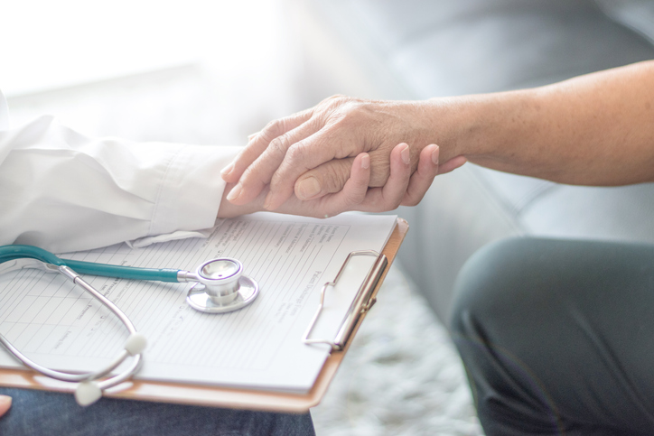 A patient holds the hand of a provider who has a clipboard on their lap.