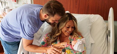 A nurse smiles while checking on a female patient and a newborn baby in a hospital bed. Family Birth Place